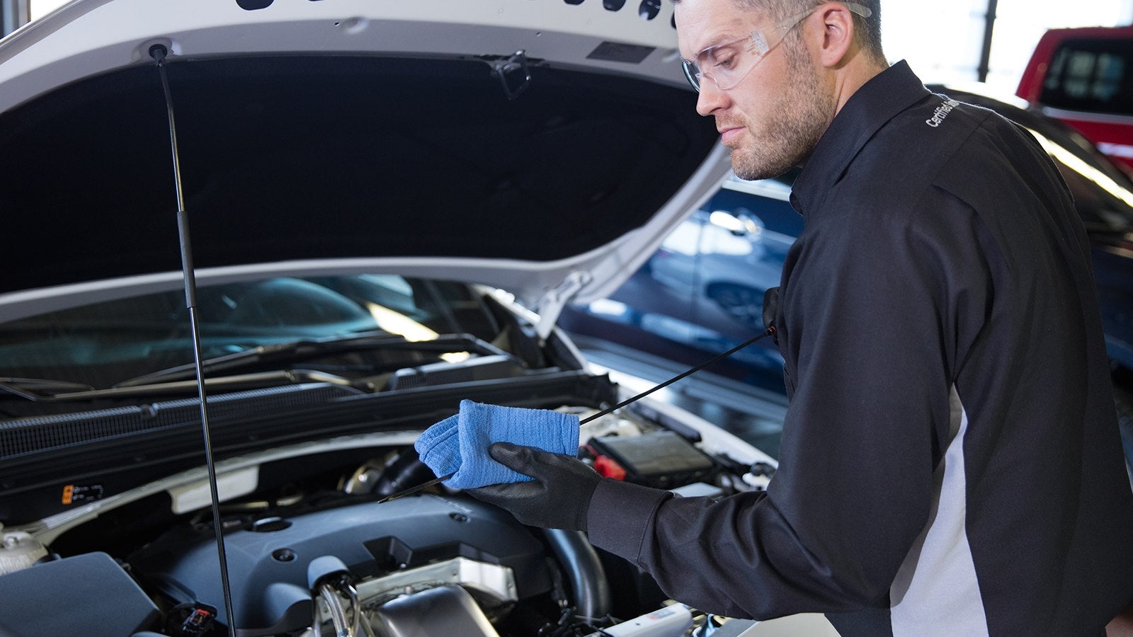 A mechanic in work attire checking under the hood of a car, holding a blue cloth and looking at the engine